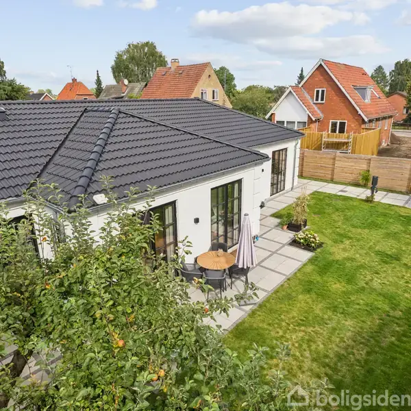 Hvidt hus med sort tegltag, placeret i en grøn have med terrasse og havemøbler. I baggrunden ses flere huse og en parkeret bil langs en vej.