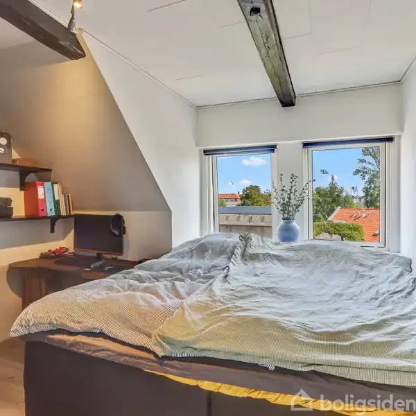 Seng med sengetøj står i et soveværelse med skrå væg. Tøj hænger på bøjler, og en hylde har bøger og papirer. To vinduer viser blå himmel og røde tage udenfor.