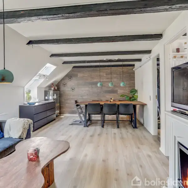 Stue med sofa, sofabord og fladskærms-tv på højre væg. Langbord med seks stole under tre lamper i baggrunden. Træbjælker og skråvinduer skaber en hyggelig atmosfære.