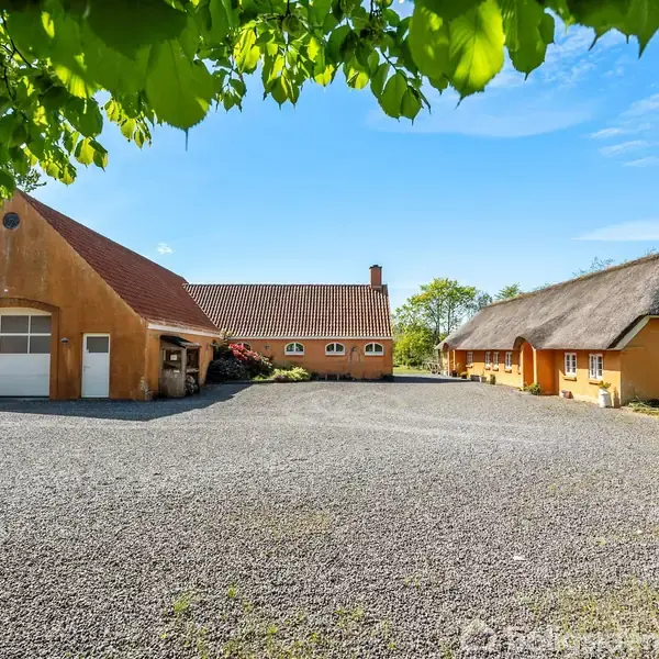 To huse med gule facader står mod hinanden på en grusbelagt gårdsplads. Det ene hus har et stråtag; det andet har en garageport. Bladfyldte grene hænger ovenfor.