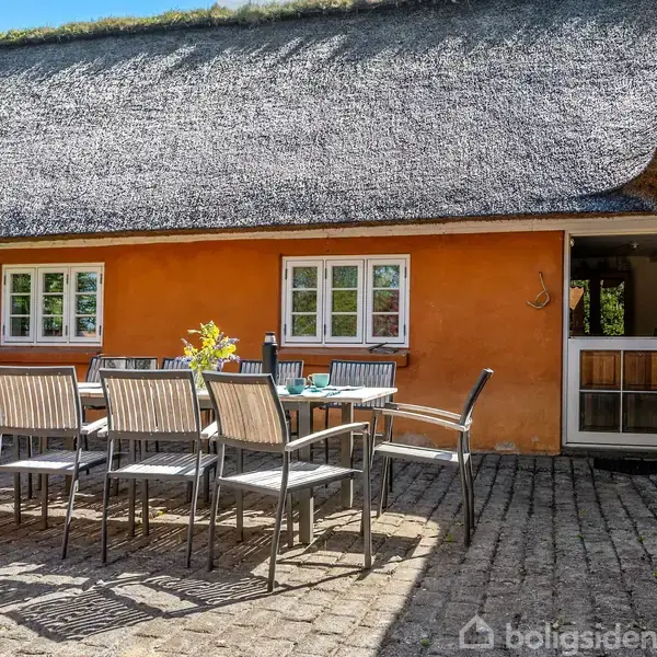 En terrasse med bord og ti stole står på et brostensbelagt areal. I baggrunden ses en gul bygning med stråtag, vinduer og en åben dør.