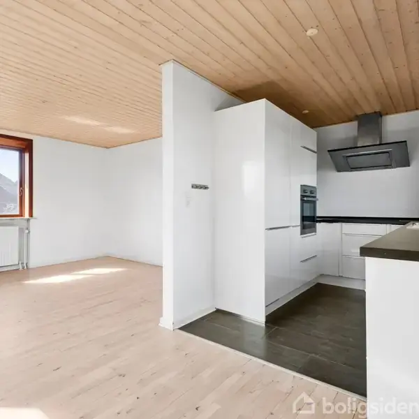 Moderne køkken med hvide skabsfronter og sort bordplade. Induktionskomfur og rustfri emhætte. Store vinduer giver naturligt lys med udsigt til en frodig have. Trægulv og træloft.