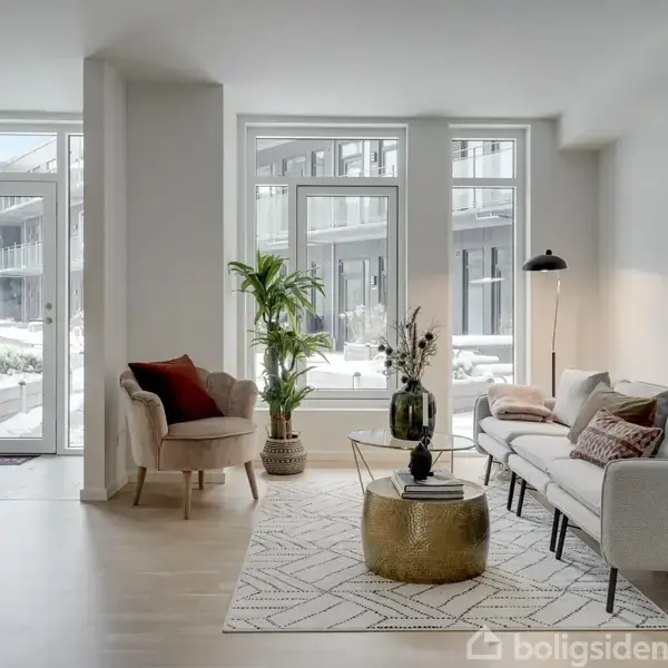 Stue med lyse møbler; sofa og lænestol er placeret omkring et rundt guldbord. Store vinduer giver udsigt til en gårdhave. En gulvlampe og planter ses også i rummet.