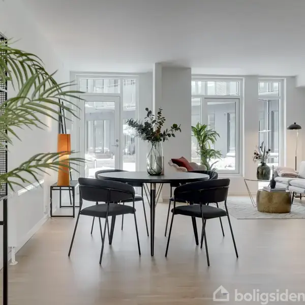 Spisebord med stole står i centrum, omgivet af lyse rum med store vinduer og planter. En sofa ses i baggrunden ved et sofabord med lamper og puder.