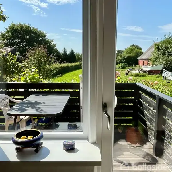 En havedør med udsigt over en terrasse med et mørkt bord og hvid stol. I baggrunden ses en grøn have med træer, en rød bygning og en trailer på græsplænen.
