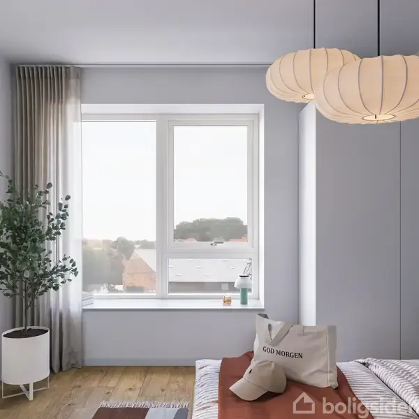 Et soveværelse med en seng, hvorpå der ligger en pude med teksten "GOD MORGEN". Ved siden af står en plante i hjørnet. En stor vindue og to lamper præger rummet.