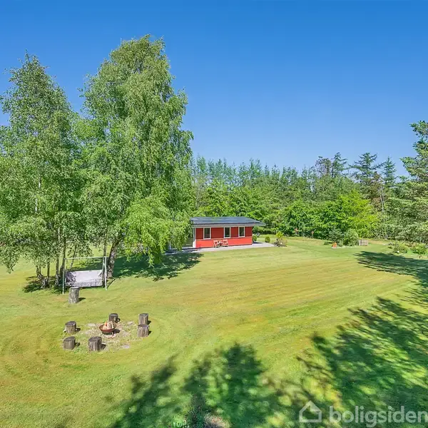Et rødt hus står stille i en grøn, skovklædt have. Ved siden af ses en ildplads omgivet af træstubbe til at sidde på. Blå himmel danner baggrunden.