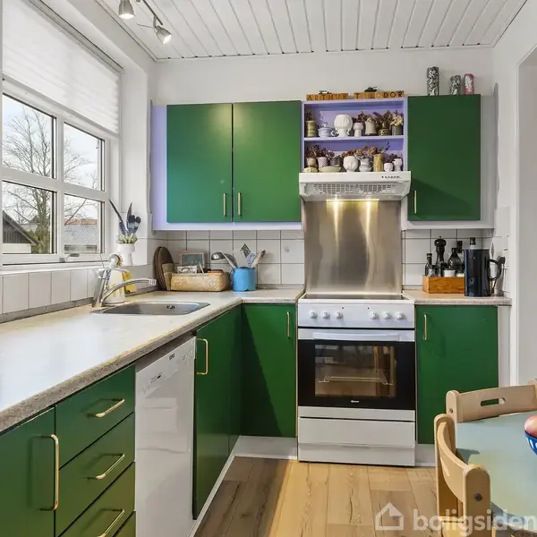 Et køkken med grønne skabe og en komfur med ovn, omgivet af hvide vægge og træbordplader. Der er en frugtskål på bordet og vinduer, der lader sollys komme ind.