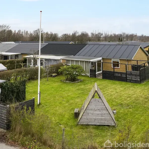 Et gult, enfamiliehus med mørkt tag står på en veltrimmet græsplæne. En flagstang er placeret i haven ved siden af en legepyramide i træ. Omkranset af hegn og buske.