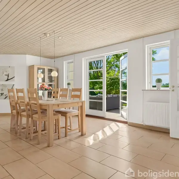 Et spisebord med seks stole står centralt i et lyst rum. Store vinduer og en dør fører ud til en terrasse med naturudsigt. Væggene har planteillustrationer.