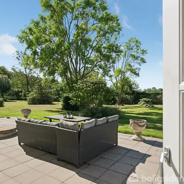 Sofagruppe står på en fliseterrasse med udsigt til en grøn have og store træer under en blå himmel. Døren til terrassen er åben, hvilket inviterer til udeliv.