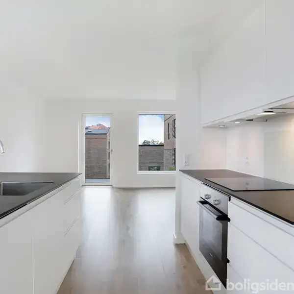 Et moderne køkken med hvidt interiør, en indbygget ovn og en køkkenø med sort bordplade og vask. Store vinduer i baggrunden giver udsigt til lysfyldt rum.