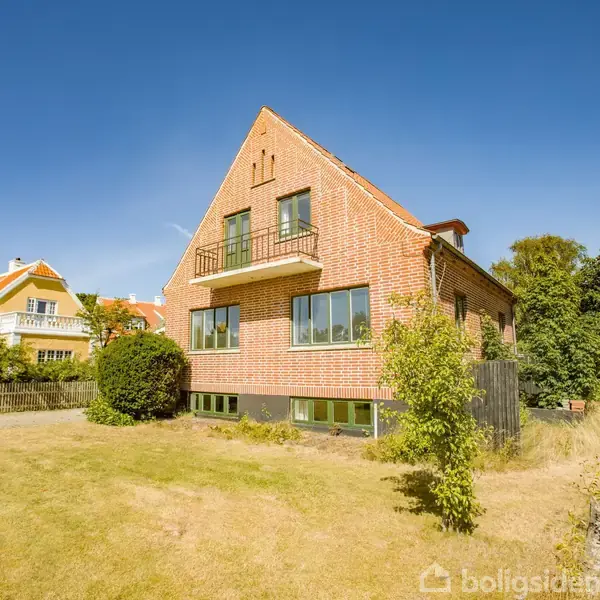 Rødstenshus med gavl og altan står i en solrig have med græsplæne og buske, omgivet af andre huse og træer under en klar blå himmel.