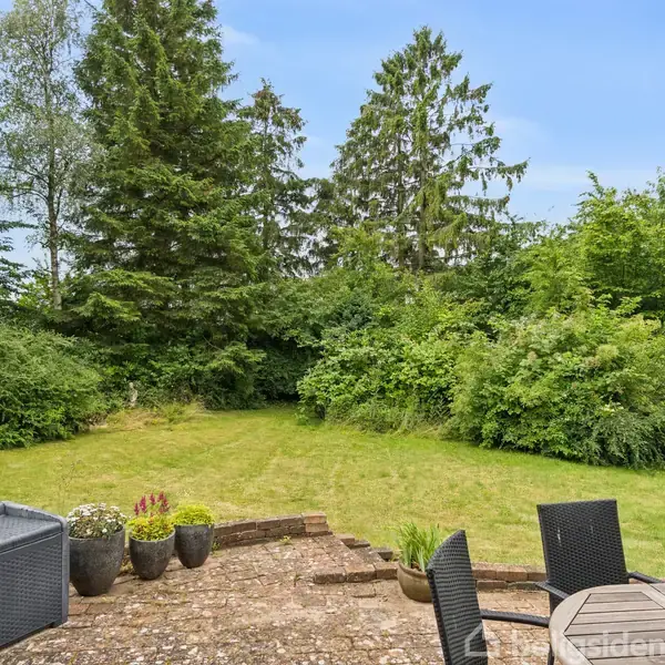 Terrassemøbler står på flisebelagt område, omgivet af flere potteplanter. En velplejet græsplæne strækker sig mod tæt bevoksede buske og høje træer i baggrunden under en klar blå himmel.