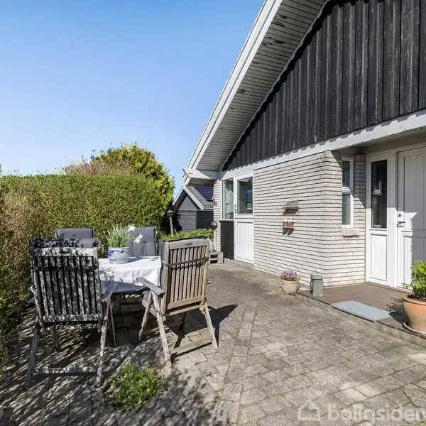 Havebord med stole står på en stenterrasse foran et hus med hvid murstensfacade. I baggrunden ses en høj hæk og klart blå himmel.