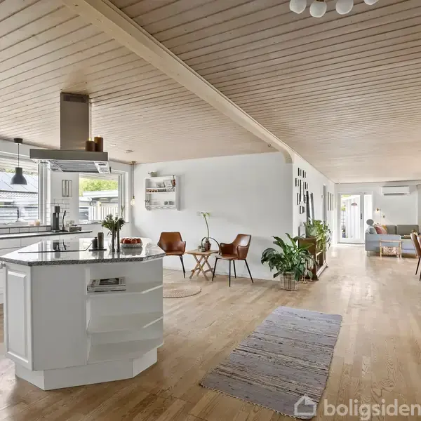 Et moderne køkkenalrum med en central ø med komfur, omgivet af hvide skabe og vinduer. Til højre ses spiseområde med træbord og stol, og i baggrunden en stue.