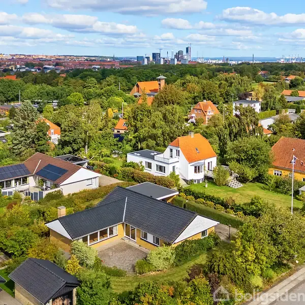 Flere huse med røde tegltage ligger stille i en grøn forstad. Omkring dem er der velholdte haver og træer; i baggrunden ses en bysilhouette med højhuse.