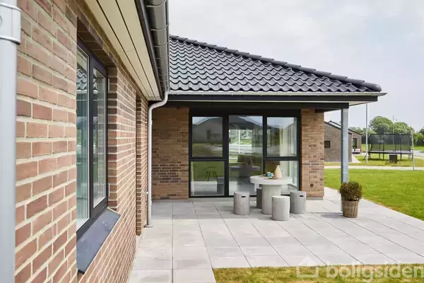 Murstenshus med sorte tagsten og store glaspartier. En terrasse med flisebelægning og moderne betonmøbler står udenfor. Græsplæne i baggrunden under en overskyet himmel.