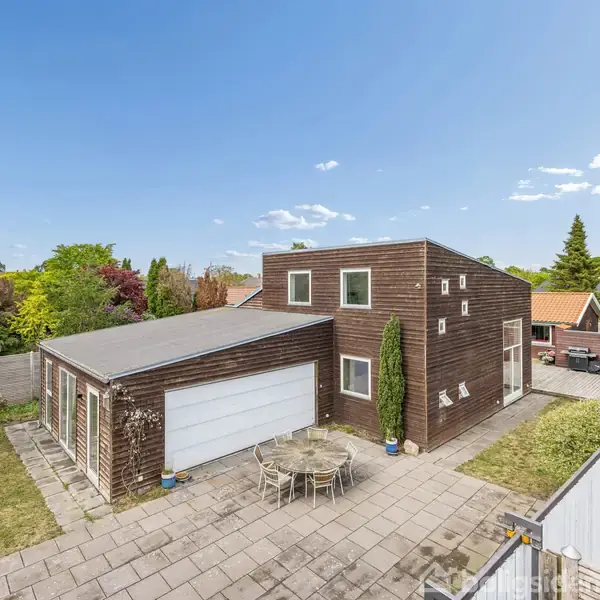 Et moderne hus med fladt tag og træbeklædning står stille på en flisebelagt terrasse omgivet af grønne arealer med skiftende vegetation, herunder enkelte hække og træer i en villakvarter.