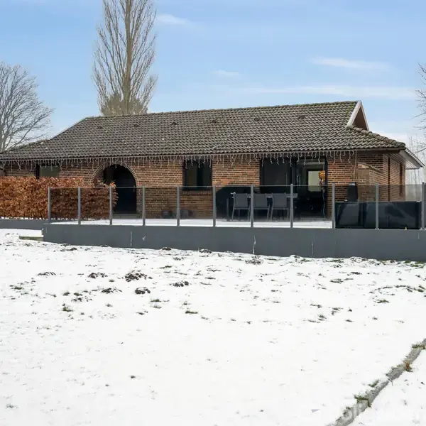 En brun murstensbungalow står stille omgivet af en snedækket have. En terrasse med glasrækværk er foran huset, mens træer hæver sig mod en klar blå himmel i baggrunden.