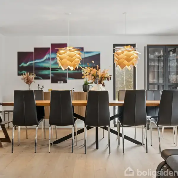 Langt spisebord med otte sorte stole står under to lamper. Bordet er dekoreret med blomster. I baggrunden hænger kunst på væggen, og et vitrineskab står tæt på.