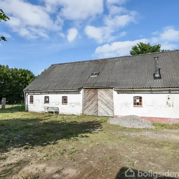 En ældre, hvid bygning med et sort tag og trægavl står på en solbelyst grund omgivet af grønne træer og buske under en blå himmel med skyer.