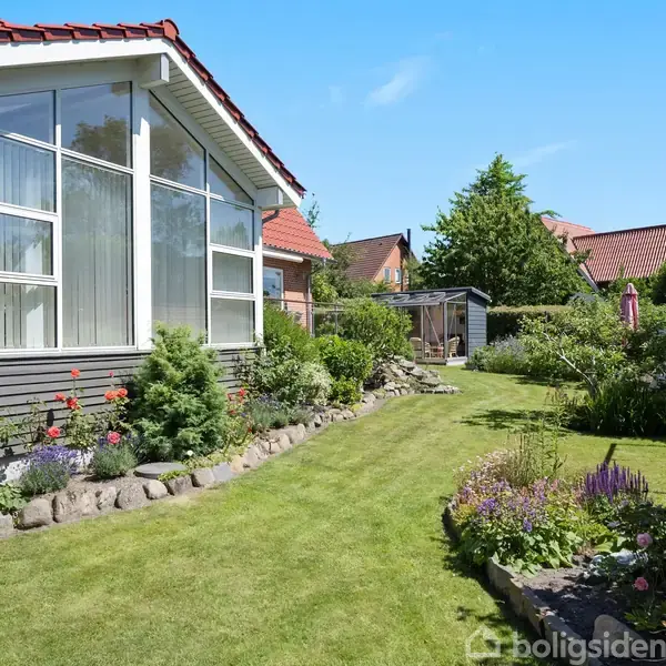 Et hus med glasparti og rødt tag står ved siden af en velplejet have med blomsterbede og græsplæne. Træer og buske omgiver området under en blå himmel.