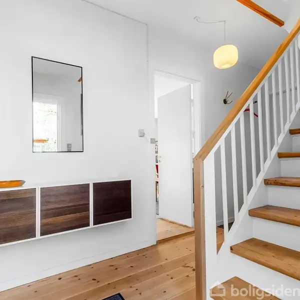 Et trætrappe fører opad i en lys entré. Væghængte skabe og et spejl hænger på den hvide væg. To runde lamper hænger fra loftet.