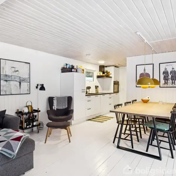 Grå sofa med tæppe står ved siden af lænestol og sofabord i en moderne, hvidmalet stue. Til højre er et spisebord med otte stole og en gul lampe.