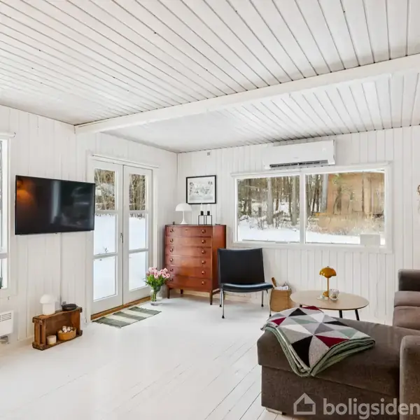 En lys stue med sofa og sofabord, hvor en blomstervase står. Et TV hænger på væggen. Store vinduer og terrassedør afslører snebelagt natur udenfor.