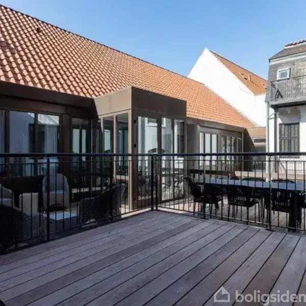 Terrasse med træplanker, møbleret med et rundt bord og kurvestole til venstre, et rektangulært bord med stole til højre, omkranset af moderne bygninger med rødt tegltag.