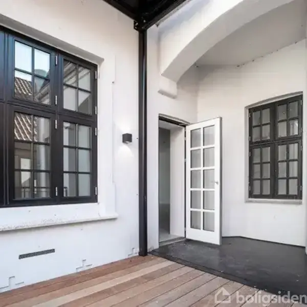Dør står åben, fører fra en træterrasse ind i et lyst rum. Store, mørke vinduer pryder væggene omgivet af en hvid facade.