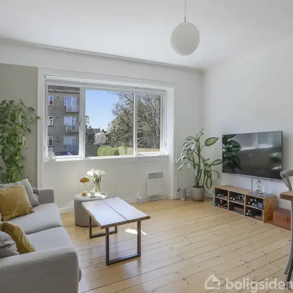 Stue med sofa og borde placeret på trægulv, planter ved vinduet. En fladskærm står på et møbel til højre. Udenfor ses en lys dag med træer og bygninger.
