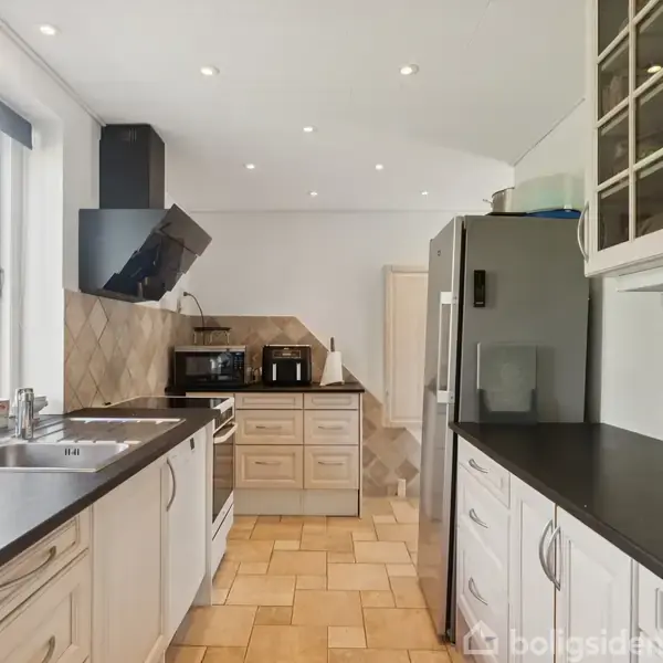 Køkken med hvide skabe og sorte bordplader. En vask står under et vindue, og der er en køleskab mod højre. Ovn og mikroovn er på afstand.