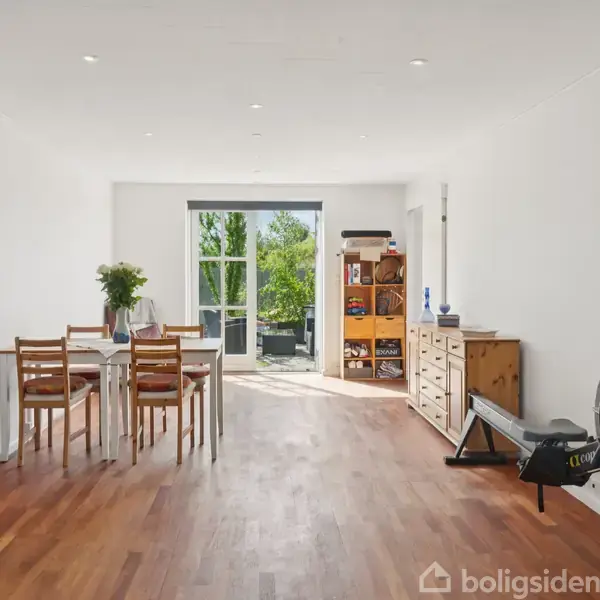 Et lyst rum med trægulv indeholder et spisebord med fire stole til venstre og en romaskine til højre. Glasdøre i baggrunden åbner ud til en balkon.