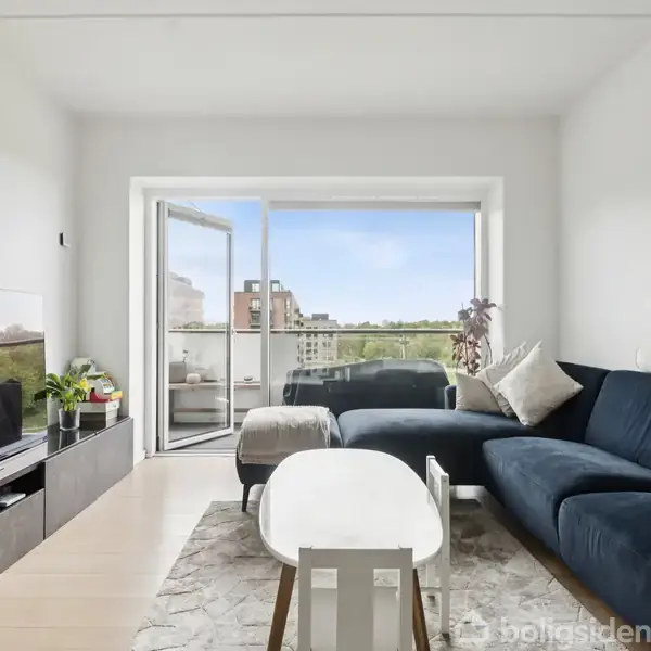 Mørkeblå sofa, stående mod hvid væg i moderne stue, foran rektangulært sofabord, store vinduer mod balkon med udsigt over bygninger og himmel, lys trægulv, fjernsyn og planter på tv-bord.