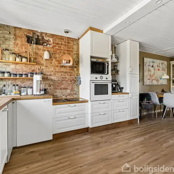 Køkken med hvide skabe og indbyggede ovne. Træhylde med krukker på murstensvæg. Spiseområde med bord og seks stole i baggrunden. Laminatgulv og loft med træpaneler.