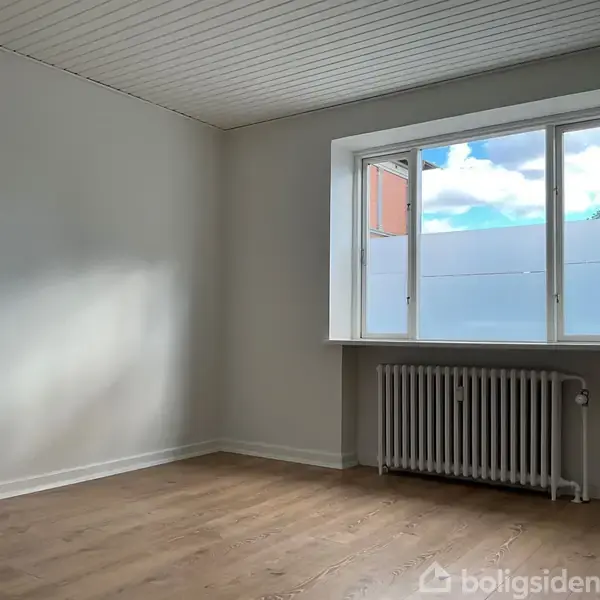 Et tomt rum med hvidmalede vægge. En radiator står under et stort vindue, der lukker naturligt lys ind. Trægulv og et hvidt loft skaber minimalismens indtryk.