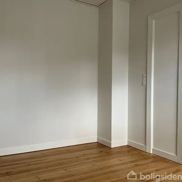 Dør står åben i et rum med lyse vægge og trælaminatgulv, hvilket skaber en ren og minimalistisk indretning.