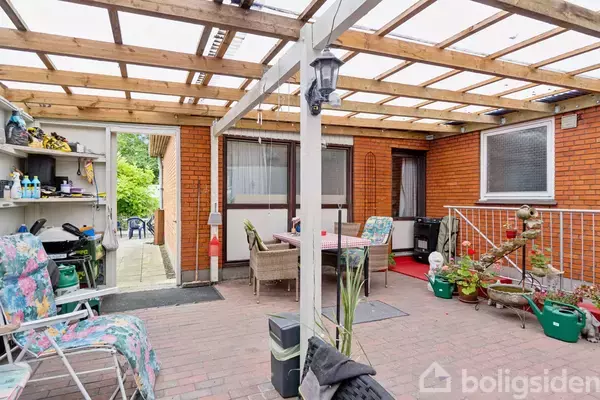 En overdækket terrasse med murstensvægge har havemøbler og planter, omgivet af en reol med forskellige genstande. En trappe fører til et indendørs område.