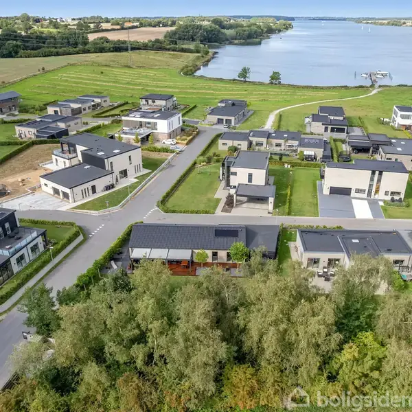 Moderne huse står stille i et nybygget boligområde omgivet af grønne marker og en bred flod, der bugter sig i horisonten.
