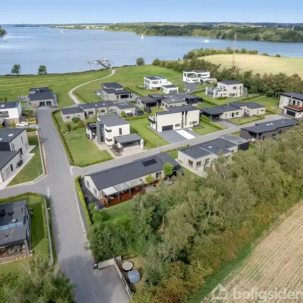 Moderne huse med flade tage ligger langs snoede veje. Omkringliggende natur omfatter træer og en stor sø i baggrunden. Landskabet er åbent og grønt med udsigt over vandet.