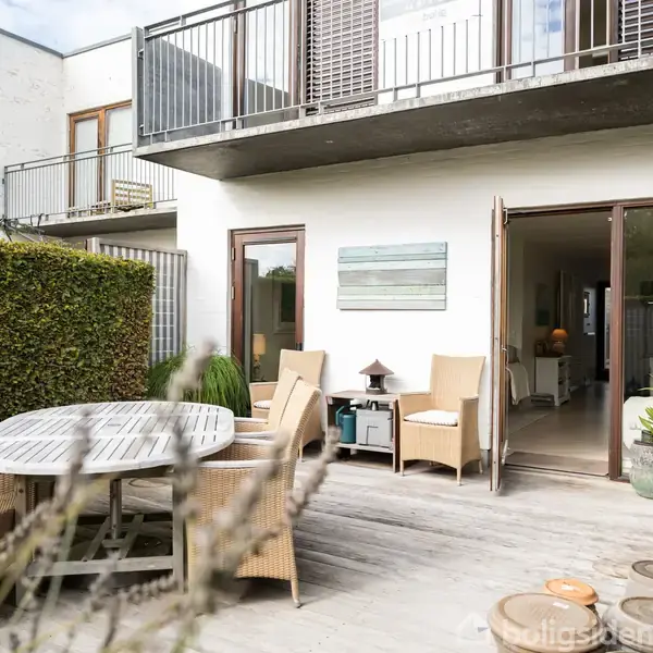 Terrasseområde med rundt træbord omgivet af stole, beliggende foran et moderne hus med åbne glasdøre. Hækken til venstre og krukker med planter skaber en hyggelig atmosfære.
