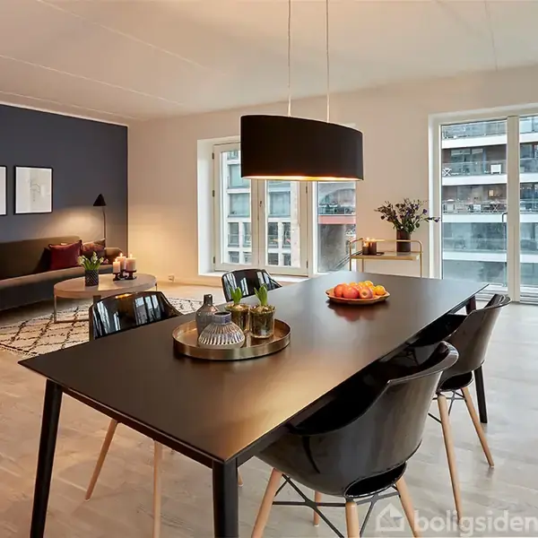 En moderne spisestue har et sort bord med frugter og glas. Omtrent fire stole omgiver bordet. Området er veloplyst med pendellampe over bordet og store vinduer med udsigt til kanaler. I baggrunden ses en sofa med puder, en tæppe og vægkunst.