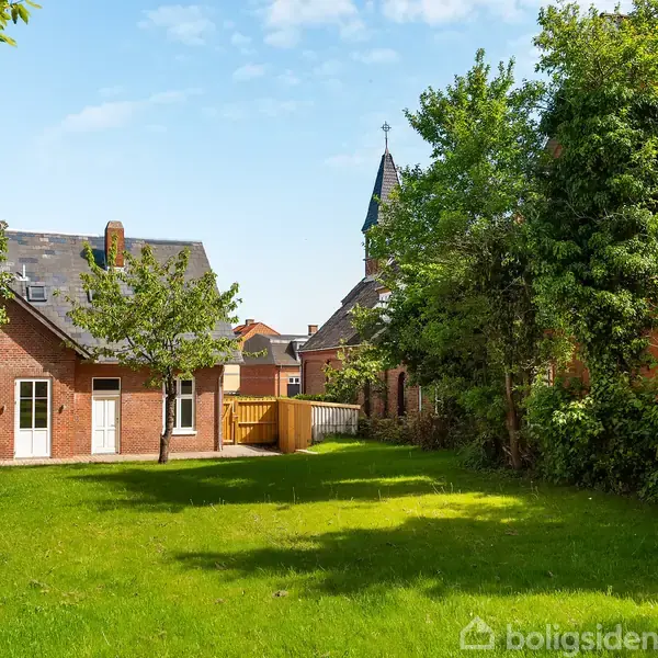 Hus med røde mursten står stille i en frodig have med grønne træer. Bag det ses en kirke med spir, omgivet af flere bygninger under en blå himmel.