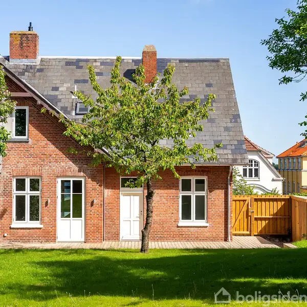 Rødt murstenshus med grå skifertag står i en solrig have. Et lille træ vokser foran huset, mens en træport leder til bagsiden, hvor flere huse ses i baggrunden.