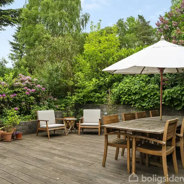 Terrasse med træmøbler og parasol. To loungestole og et lille bord står til venstre, omgivet af frodig grøn beplantning og blomster. En rolig udendørs atmosfære i en have.