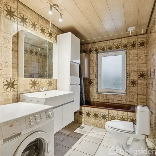 Badeværelse med brune fliser og mønster, vaskemaskine ved siden af håndvasken, badekar ved et vindue, og toilet i højre hjørne. Loftet er panelbeklædt. Ingen tekst.