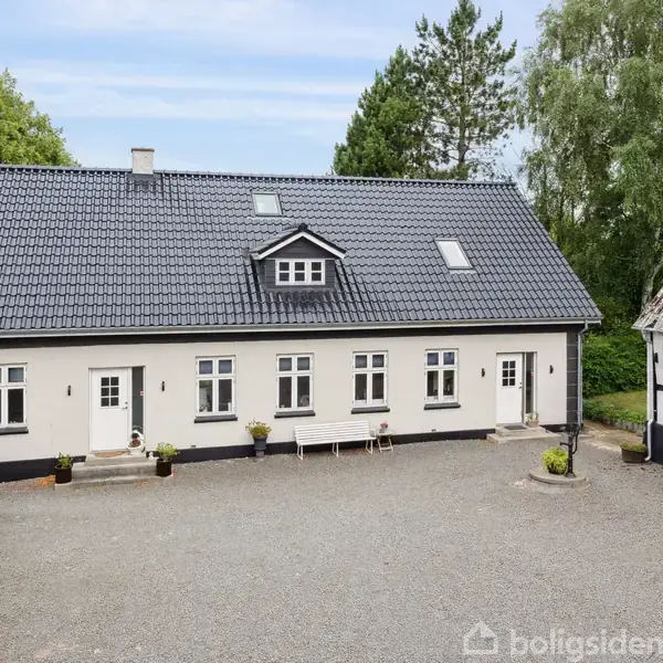 Et hvidt hus med gråt tag står på en grusbelagt gårdsplads. Flere vinduer ses langs facaden. Omgivet af træer og et ældre udhus til højre.