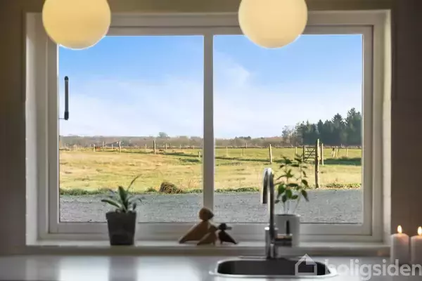 Køkkenvindue med udsigt over grønne marker og fjerne træer; to kugleformede lamper hænger ned fra loftet, og planter står på vindueskarmen ved siden af en vandhane.
